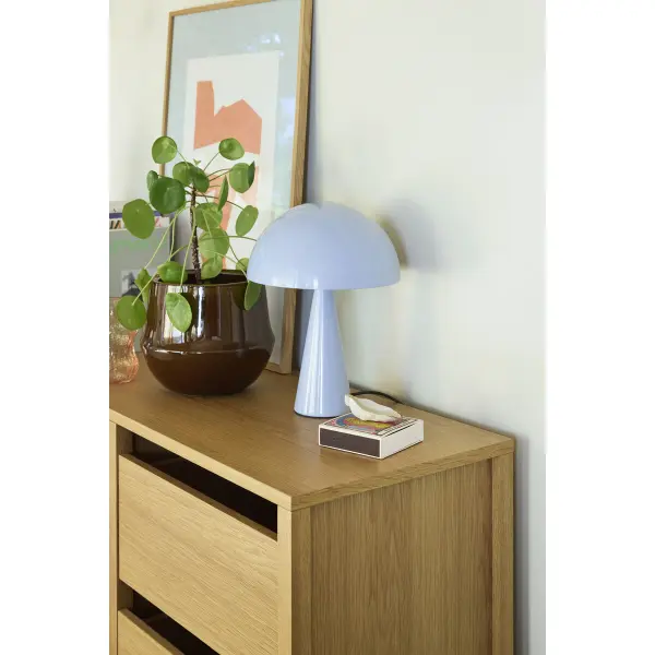 Lampe de table bleu glacier au design minimaliste posée sur un meuble en bois clair, diffusant une lumière chaude et apaisante dans un salon.
