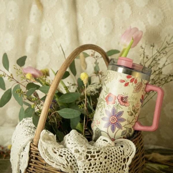 Mug isotherme XL de 1,2L avec motifs floraux bohèmes, posé dans un panier en osier garni de fleurs et d'un napperon en dentelle.