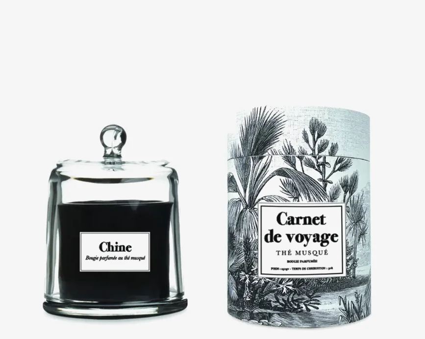 Une bougie noire dans un bocal en verre avec couvercle cloche, à côté de son emballage cylindrique avec une étiquette 'Carnet de voyage'.