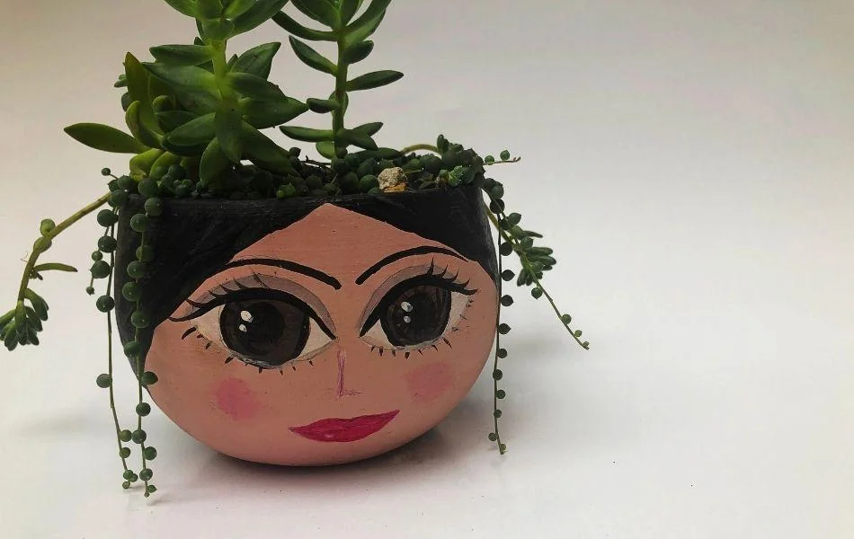 Pot en terre cuite peint à la main représentant un visage de femme avec une plante succulente suspendue en guise de cheveux.