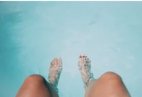 Vue subjective des jambes et des pieds plongés dans l'eau bleu clair d'une piscine en été.