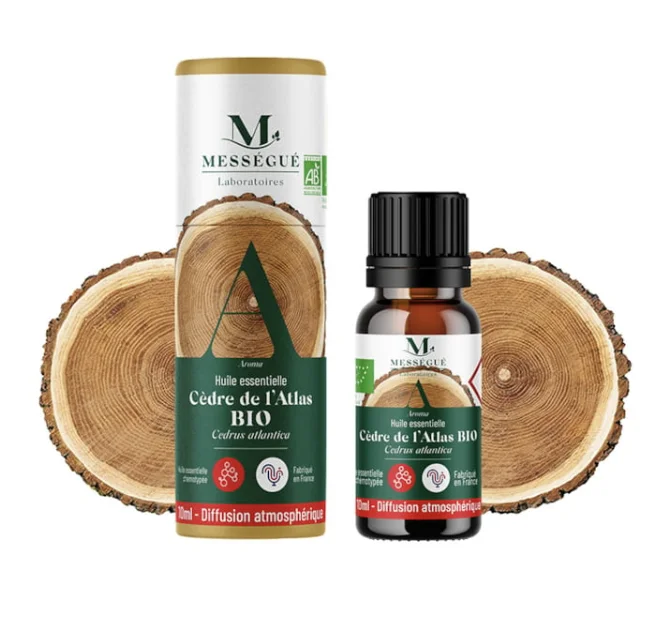 Flacon d'huile essentielle de Cèdre de l'Atlas BIO Mességué de 10ml à côté de son emballage tubulaire, devant deux rondins de bois texturés.