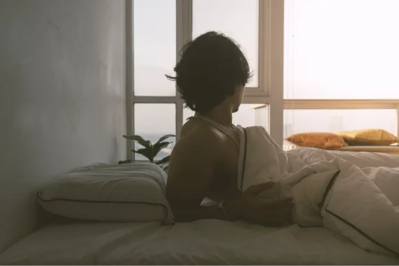 Une personne assise dans son lit regardant par la fenêtre sous la douce lumière du matin.